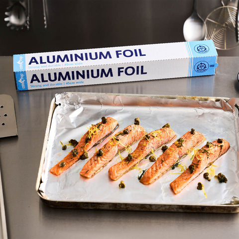 A box of Calphite aluminium foil sits on a stainless steel countertop behind a baking tray lined with foil and holding cooked salmon fillets topped with herbs and lemon zest.